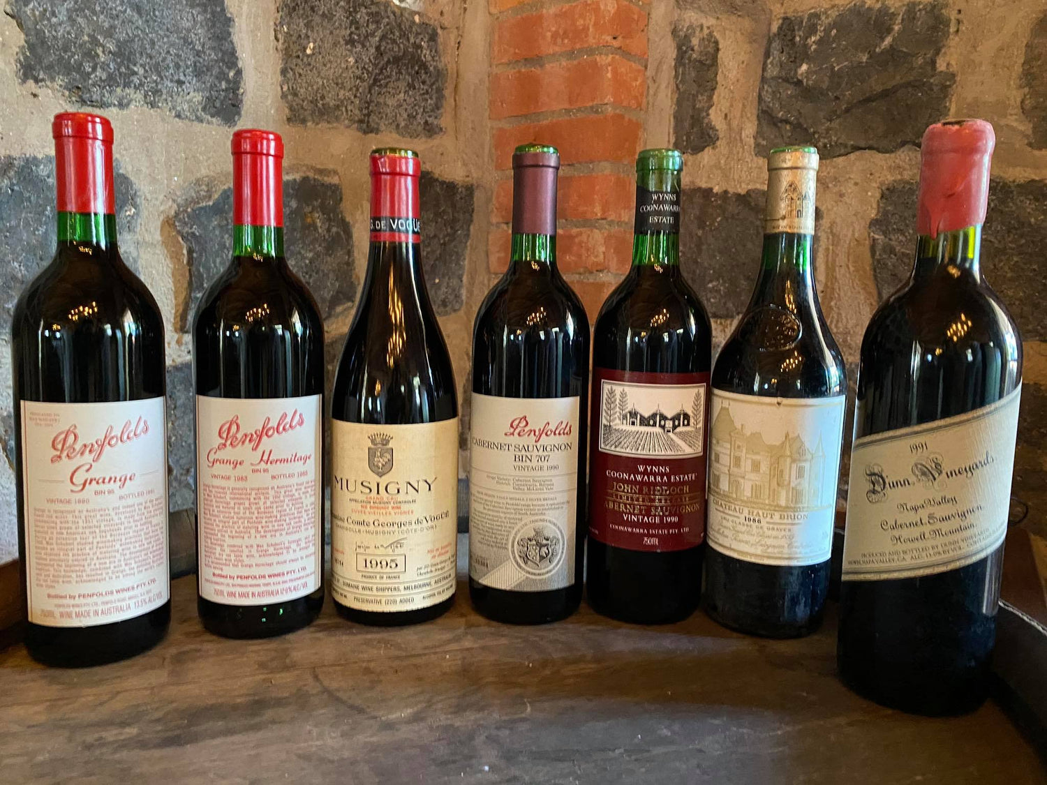 Seven bottles of wine on top of a wine barrel against the Bluestone Cottage wall.